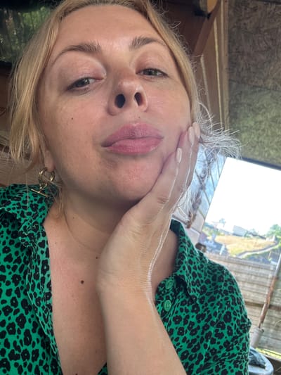 In Razdolnoye, Russia, a woman with blonde hair and light skin is captured in a close-up, selfie-style shot. Her right hand is gently placed on her face, with her fingers resting on her cheek. She is wearing a green shirt with a black leopard print. Her lips are slightly parted and painted with pink lipstick. A gold hoop earring with a bar detail is visible on her left ear. The background suggests an outdoor setting, possibly a rustic or unfinished structure, with a glimpse of a bright sky and a distant landscape.