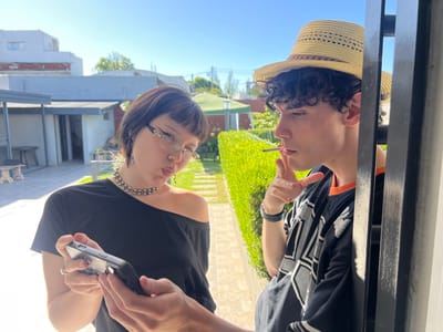 The image depicts two individuals in a residential outdoor setting in Quilmes Oeste, Argentina, during daylight hours under a clear blue sky.
On the left, a female subject with short dark hair and eyeglasses wears a black off-the-shoulder top and a studded choker necklace. She holds a black smartphone horizontally with both hands, looking down at the screen with her lips pursed in a pout. A ring is visible on the ring finger of her right hand.
To her right, a male subject with short curly dark hair wears a light-colored straw hat and a dark jersey with orange trim and white graphics. He stands partially obscured by a black vertical metal bar, leaning into the frame. He has a hand-rolled cigarette in his mouth, which he is actively smoking, and his right hand is raised to chest level. He looks towards the left, appearing to observe the female or the smartphone.
The background features a paved patio area on the left, transitioning into a green lawn or garden path in the center. A dense, trimmed green hedge borders the right side of the garden path. Multiple residential buildings are visible in the distance beneath the clear sky, indicating a suburban environment.