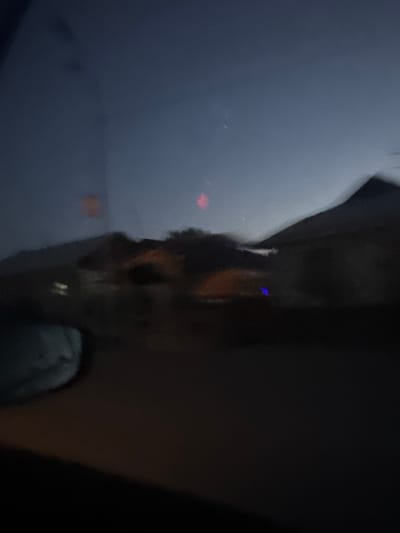 The image depicts a blurry, low-light outdoor scene, likely taken from inside a vehicle, possibly a car given the visible side mirror on the left. The time of day appears to be dusk or night, with a dark blue sky. The background shows indistinct shapes of buildings with roofs, suggesting a residential area. There are scattered lights, some appearing as blurred orange and blue glows, possibly from streetlights or illuminated windows. The overall impression is one of movement, with the lack of sharp focus indicating the photograph was taken while the camera was in motion. No people are clearly discernible in the image. The location is identified as Keŋ-Say, Kyrgyzstan, which suggests this is a view of a neighborhood in that city at night.
