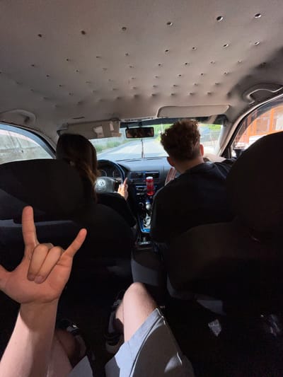 The image shows the interior of a car with two people. A person in the foreground on the left is holding up a hand with fingers forming the "rock on" or "devil horns" gesture. This person's legs, wearing gray shorts and black and white sneakers, are also visible. In the driver's seat is a person with curly brown hair, facing forward and appearing to be driving. A red Coca-Cola can is visible in the center console. A passenger with long dark hair is in the passenger seat, facing forward. The car's ceiling has numerous small indentations. The scene appears to be on a road, possibly in a rural or suburban area, with some buildings visible in the background through the windows. The specific location in Vlahovo, Bulgaria is not discernible from the visual information provided.