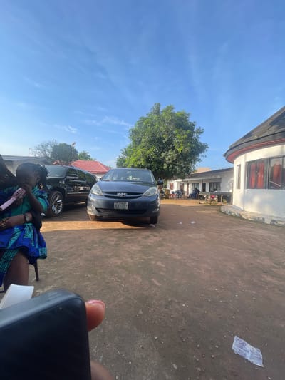In Orodo, Nigeria, a scene unfolds under a clear blue sky with wispy clouds. The foreground features a person's hand holding a black rectangular electronic device, possibly a phone or power bank, obscuring a portion of the bottom left of the image. To the left, a child, wearing a vibrant blue and green patterned outfit, sits holding a pink toy microphone. Behind the child, a black SUV is parked. In the center of the frame, a dark blue minivan is parked on a dirt road. Further back, several buildings are visible, including a white structure with a circular section and red-curtained windows, and another building with a red-tiled roof. Trees with lush green foliage are scattered throughout the scene. Some individuals are seated at tables in front of one of the buildings, suggesting a gathering or social event. The ground is a mix of dirt and gravel, with a few pieces of trash scattered about.