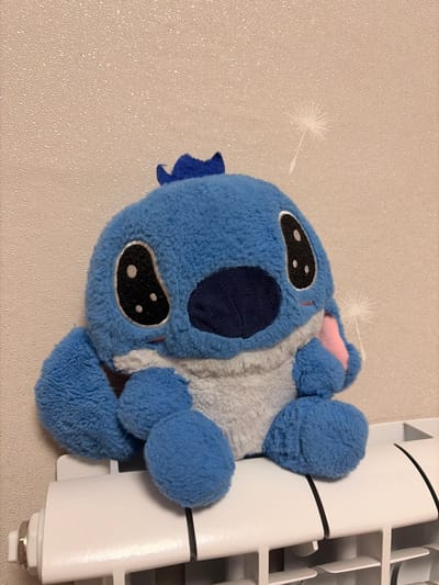 A blue plush toy, resembling the character Stitch, sits on a white radiator against a textured, light-brown wall. The toy has large black eyes with white pupils, a dark blue snout, and pink accents on its ears. A small blue tuft is on its head. The wall has a subtle pattern and a faint, faint white dandelion-like silhouette. The radiator appears to be in a domestic setting, possibly a home in Leninogorsk, Russia, though no specific city or country identifiers are visible.