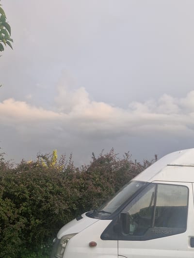 The image depicts a white Ford Transit van parked outdoors, with a dense hedge of foliage behind it. The sky above is overcast with soft, diffused light, suggesting it may be late afternoon or early morning. The van's exterior shows signs of recent rain, with water droplets visible on the windows and body. A portion of a green leafy branch hangs into the frame from the top left corner. The setting appears to be a rural or suburban area, possibly within Saulgoži, Latvia, given the context. There are no people visible in the image.