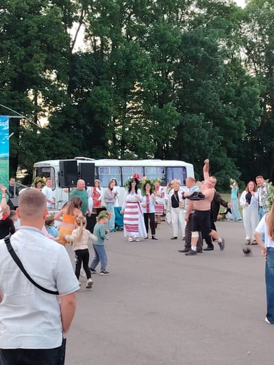 The image depicts an outdoor gathering in front of a white bus, possibly at a cultural event or festival in Олтушскі сельскі Савет, Belarus. A group of people are present, with many wearing traditional attire, including floral wreaths on their heads. In the foreground, a man in a white shirt and black pants faces away from the camera, with a black strap from a bag draped over his shoulder. To his left, children are visible. In the center of the scene, a man without a shirt, wearing black shorts, is in mid-action, possibly performing a feat of strength or participating in a game. Other individuals are observing the activities, with some clapping or reacting to the events. The background features a dense line of tall green trees, suggesting the event is taking place in a semi-rural or park-like setting. The ground is paved, indicating an outdoor public space. The lighting suggests it is daytime, with the sun possibly setting or being partially obscured by the trees.