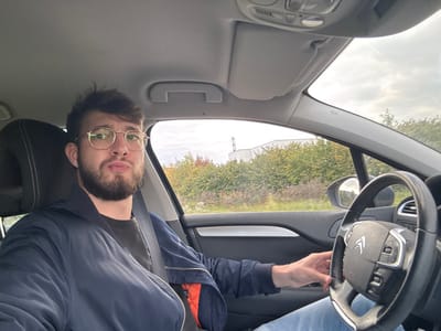 A male individual is depicted seated in the driver's seat of a Citroën vehicle, located in Scherwiller, France. The subject has short dark hair, a beard, and wears round gold-rimmed eyeglasses. He is dressed in a dark blue bomber jacket over a dark t-shirt and blue jeans, with a seatbelt visible across his chest. His right hand rests on the steering wheel. Through the vehicle's windows, an outdoor environment is visible, featuring green and autumnal foliage, a large light-colored industrial building with a cylindrical structure in the distance, and a cloudy sky. The car's interior features a light-colored headliner and a dark dashboard and door panel.