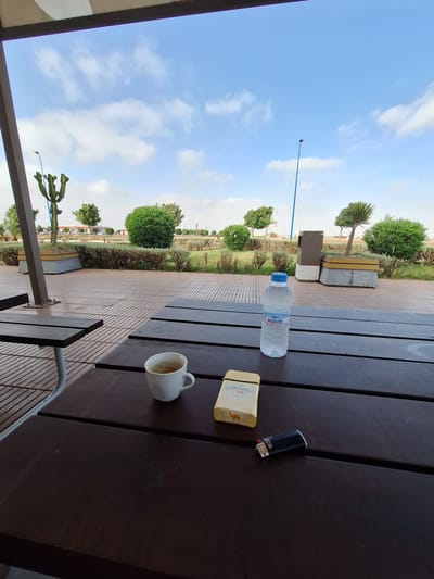 A low-angle shot shows a dark wooden picnic table with several items on it, set against a backdrop of a park-like area with lush green bushes, trees, and a bright blue sky with scattered white clouds. In the foreground, a white ceramic mug filled with a dark liquid, likely coffee, sits on the left side of the table. To its right, a yellow pack of Camel cigarettes is positioned next to a black lighter. A clear plastic bottle of Sidi Ali brand water stands upright on the table to the right of the cigarette pack. The table is partially covered by an overhead structure with a brown and white striped awning. In the background, a light blue lamppost extends upwards, and beyond that, a street and buildings are visible. The scene appears to be outdoors, possibly at a rest stop or public park in Oulad Hcine, Morocco, judging by the Sidi Ali water brand. There are no people visible in the image.