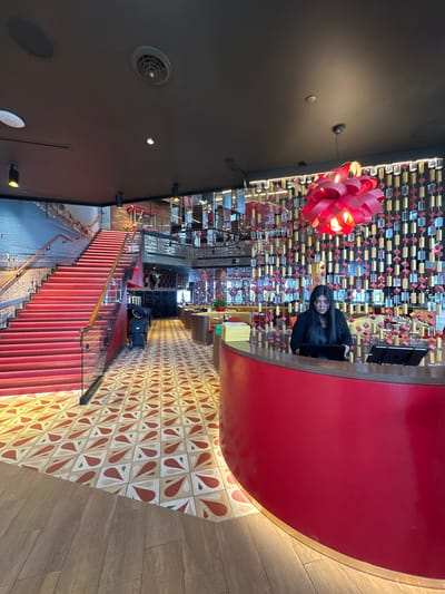 The image depicts an interior space in Lake Buena Vista, United States. A person with long dark hair is seated at a high red counter. Above them, a decorative display of small square objects hangs from the ceiling, illuminated by a red floral light fixture. Stairs lead downwards, adjacent to a wall displaying a mural of a beach scene. The floor is tiled with a geometric pattern, transitioning to a wooden plank design. Luggage is visible further down the corridor. The ceiling above is dark with visible light fixtures and a ventilation grate.
