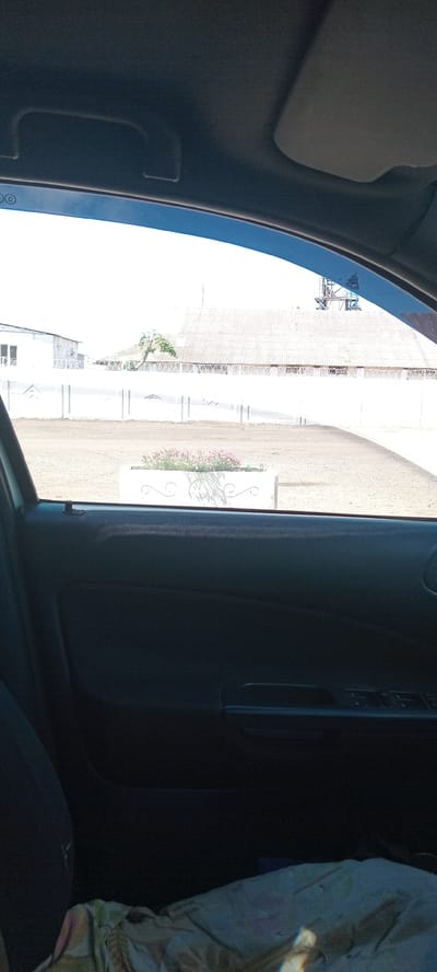 The image shows a view from inside a car looking out through the open window. The exterior scene features a white wall with a decorative pattern and barbed wire along the top, a white building with windows in the background, a white corrugated roof structure behind the wall, and some industrial equipment visible above the roofline. In the foreground of the exterior, there is a rectangular white planter box containing pink flowers. The interior of the car shows a dark-colored car door panel with window control buttons. A fabric-covered object is visible on the car seat in the lower portion of the image. The sky is bright and appears to be daytime. The location is specified as Koviahy, Ukraine.
