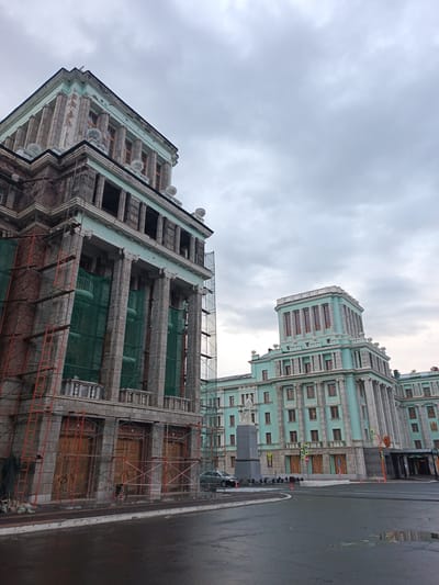This media file captures a low-angle view of a grand, teal-colored building with neoclassical architecture, likely undergoing renovation. Scaffolding, constructed from bright orange metal poles and covered with green netting, encases the left side of the building. Large wooden doors with visible bracing are present on the ground floor, suggesting active construction or a protected facade.
The building exhibits a series of tall, fluted columns on its upper levels and decorative cornices. The sky above is overcast with heavy, grey clouds, indicating a gloomy or possibly rainy day. The street in front of the building is wet and reflective, with a large puddle in the foreground, further emphasizing the damp weather.
To the right, a continuation of the teal building is visible, featuring rows of windows and a similar architectural style. A statue stands on a pedestal in front of this section of the building, facing the street. Yellow pedestrian crossing signs and traffic lights are also visible, indicating an urban environment. The overall mood is somber and perhaps a bit melancholic, amplified by the cloudy sky and wet surroundings, suggesting a sense of ongoing work and a pause in activity due to the weather. The location is identified as Norilsk, Russia.