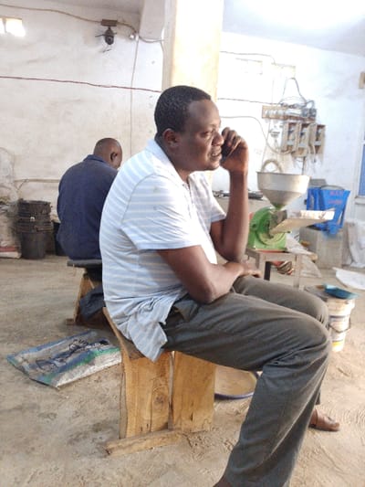 The scene is set indoors in Olomoda, Nigeria, within what appears to be a processing or workshop environment. Two adult males are present. In the foreground, a dark-skinned male, wearing a light blue and white striped short-sleeve polo shirt and dark gray trousers, sits on a rudimentary wooden bench. He is actively holding a dark-colored mobile phone to his right ear, engaged in a conversation. His left arm is bent, with his hand resting on his left thigh. He wears dark sandals. Behind him and slightly to the left, a second dark-skinned male, bald and wearing a dark blue short-sleeve shirt, sits facing away from the camera on a similar wooden bench.
The background consists of light-colored, possibly off-white, walls displaying visible wear and exposed electrical wiring. Multiple white electrical boxes and switches are mounted on the wall in the mid-ground. To the right, a green and silver machine, identifiable as a grinder or mill with a funnel-shaped hopper, rests on a small wooden table. A blue plastic container is positioned behind this machine, and a white bucket with a handle is on the floor nearby. On the left side of the image, several black buckets are stacked, and white sacks are partially visible. The floor is made of concrete, and a blue and white woven mat or bag lies on it near the foreground male's feet.