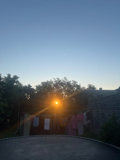 In Zviahel, Ukraine, a backyard scene unfolds under a clear, pale blue sky transitioning into twilight. The sun, a bright orange orb with visible rays, is low on the horizon, partially obscured by the dark green foliage of trees that form a dense canopy across the middle ground. A clothesline, strung between supports, is laden with various articles of laundry, predominantly light-colored garments such as shirts and dresses, some appearing to be pink and light gray. To the right, a textured grey brick wall extends into the background, its construction suggesting a boundary or part of a building. In the foreground, the rounded edge of a trampoline is visible, its dark, taut surface occupying the lower portion of the frame. The overall impression is a quiet, domestic moment captured at dusk.
