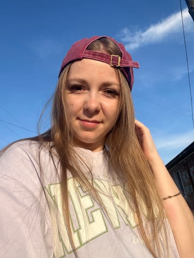 The image shows a young woman wearing a maroon baseball cap and a light purple t-shirt with green lettering. She is outdoors, with a bright blue sky in the background. The sun is casting a warm light on her face, and her hair is long and blonde. She is smiling and touching her hair with her right hand. Her left arm is visible, and she is wearing a delicate gold bracelet. Behind her, there is a hint of a building with a brick facade and a dark roofline. The scene suggests a casual, sunny day. The image was taken in the city of Mal'tsevo, Russia.