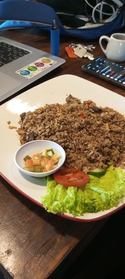 A white plate with fried rice, lettuce, and sliced tomato sits on a wooden table. A small white bowl contains pickled vegetables. Nearby are a laptop, a blue vape pen, keys, a small white cup, and a smartphone. The background includes a blue bag and tangled white cords.