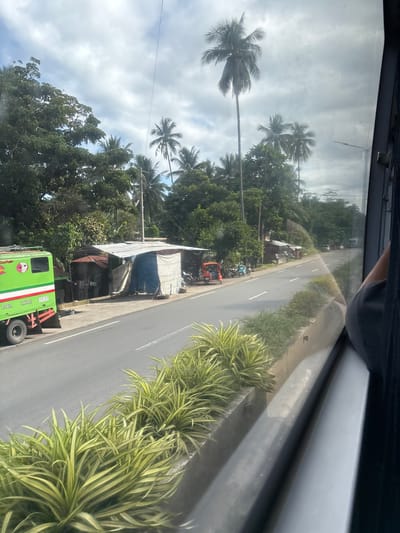 This is a photograph taken from inside a vehicle, looking out the window at a roadside scene in Santa Cruz, Philippines. The road is asphalt with white lane markings. On the left side of the road, there is a green truck with "JCA-871" license plate and a logo on its side. Adjacent to the truck are several small, makeshift structures with corrugated metal roofs and tarp coverings, appearing to be shops or dwellings. A red tricycle and several motorcycles are parked in front of these structures. Lush green trees and tall palm trees line the background, with a partly cloudy sky above. In the foreground, a row of neatly trimmed green and yellow plants lines a concrete barrier separating the road from the vegetation. The perspective suggests the vehicle is moving, as indicated by the slight blur of the scenery and the reflection on the window.