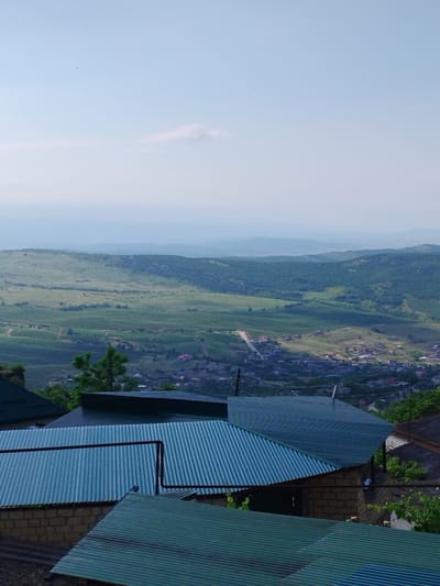 This image captures a panoramic view of the city of Darvag, Russia, from an elevated perspective. In the foreground, several corrugated metal roofs of residential buildings dominate the lower portion of the frame. These roofs are a mix of teal and dark green colors, with visible vertical ridges. The walls of some of these structures appear to be constructed from brick.
Beyond the immediate rooftops, the landscape unfolds into rolling hills covered in lush green vegetation. The mid-ground features a town or village with scattered buildings, some with red-tiled roofs, nestled within the hills. A road winds through this area. Further in the distance, layers of hazy blue mountains recede into the horizon under a pale blue sky with a few wispy clouds. A solitary dark object, possibly a bird, is visible in the upper sky. The overall impression is a serene rural landscape on a clear day.