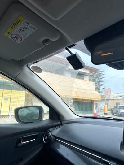 The image depicts a street scene in Latsia, Cyprus, viewed from the interior of a vehicle, specifically from the passenger side looking forward and to the right.
In the immediate foreground, the passenger-side sun visor is visible, featuring a yellow "AIRBAG" warning label with safety pictograms. A black dash camera is mounted to the windshield near the rearview mirror, which is partially visible on the right. The windshield exhibits dust and a circular sticker (likely a vehicle permit or inspection decal). The vehicle's dashboard and the upper part of the passenger door panel are visible, featuring a chrome door handle and a circular air vent.
The exterior scene reveals a two-story building across the street. Its ground floor is characterized by dark green roll-down shutters covered by a retractable yellow awning. The upper floor appears to have a light-colored facade and a reddish-brown tiled roof. To the far left, a bright yellow corrugated metal structure is present. Further down the street, scaffolding is erected against a building, indicating construction or renovation.
Street furniture includes a red and white traffic cone on the pavement, a traditional yellow pillar post box, and a signpost with a red, rectangular sign, possibly indicating a "TAXI RANK." A dark-colored SUV is parked in the distance on the right, beyond the pillar box. The sky above is overcast. No individuals are clearly identifiable or engaged in direct actions or interactions within the frame.