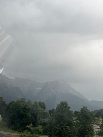 This is a photograph taken from a window, possibly a train or car, looking out at a mountainous landscape under a cloudy sky. In the foreground, lush green trees dominate the lower portion of the image, with a road visible to the left. Beyond the trees, a range of mountains rises, their peaks partially covered in snow and shrouded in mist. The sky is overcast with heavy clouds, suggesting a possibly rainy or stormy day. A very small section of buildings and construction equipment can be seen in the far right distance. The overall mood is atmospheric and somewhat somber due to the weather conditions. The image is likely taken in or near Farchant, Germany, given the presence of the Bavarian Alps. There are no people or animals visible in the frame.