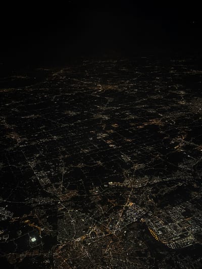 The image shows a nighttime aerial view of the city of Montegrotto Terme, Italy. The city is illuminated by a vast array of streetlights and other lights from buildings. The lights create a dense grid pattern across the landscape, with many roads and highways visible. The scene is framed by a dark, starry night sky. The image was taken at an altitude high enough to see the entire city in a single frame. The dark background, and many small lights, give an impression of vastness and isolation, despite the dense collection of lights.