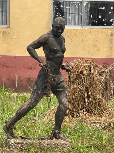 An outdoor scene in Umunekwu, Nigeria, captures a dark-colored, full-body statue of a male figure positioned in a dynamic, forward-striding pose. The statue is bare-chested and wears shorts; its wrists and ankles are secured by metal shackles connected by chains. The figure's head is bowed, directing its gaze downwards. It stands on a rectangular base composed of aggregate material. The foreground and mid-ground are covered in green grass of varying heights. To the right of the statue, a significant mound of dried, light-brown vegetation, resembling straw or hay, is present. In the background, a building features a lower wall section painted dark red and an upper wall section painted a faded yellow. Two windows with aluminum frames and security bars are visible in the upper wall, reflecting distorted white shapes.