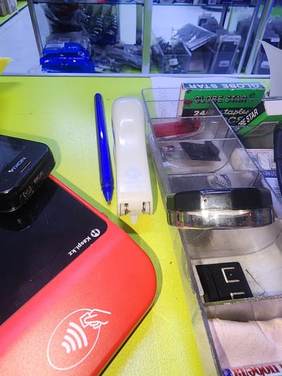 A close-up, eye-level shot captures a collection of various objects arranged on a bright yellow surface. In the foreground, a red payment terminal with the "Kaspi.kz" logo and an NFC symbol is prominent. To its left, a black Nokia phone is positioned. A blue ballpoint pen lies horizontally across the scene. To the right of the pen, a white stapler is visible. Further to the right, a clear plastic organizer holds several items, including a small metal tin with a black lid, what appear to be staples, and a small black tool. In the background, a glass display cabinet is filled with various electronic devices and accessories, suggesting a retail environment, possibly a mobile phone shop or electronics store. The overall impression is one of a cluttered desk or counter space within a commercial establishment.