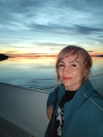 A woman with short, blonde hair and light skin is captured in a selfie against a backdrop of a sunset over a body of water. She is wearing a dark t-shirt with a graphic print and a teal jacket. The sky is a gradient of blues and oranges, with wispy clouds. The water reflects the colors of the sky and shows ripples from movement. The foreground shows a light-colored railing or deck of a vessel. The woman is smiling and looking directly at the camera. The scene suggests a tranquil evening, possibly on a lake or river near Sheksna, Russia, given the terrain visible on the horizon and the type of sky and water.