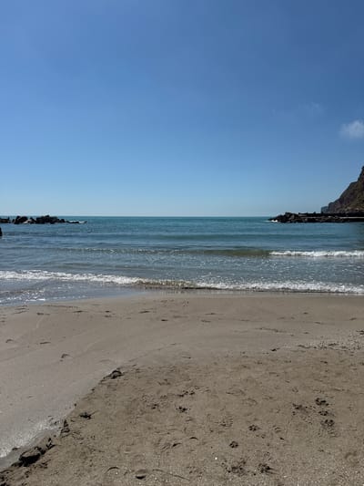 The image depicts a beach scene likely in or near Balgarevo, Bulgaria. A stretch of sandy beach occupies the right side of the frame, marked with footprints. The sea lies adjacent to the beach, with small waves visible. A rocky structure borders the sea. The left side of the frame features a clear blue sky with minimal clouds.