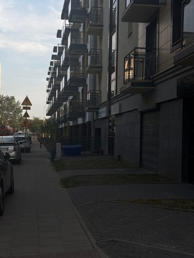 The image depicts a street scene in the city of Białystok, Poland. On the right side of the frame, a modern multi-story building with balconies is visible. The building's facade is a mix of light and dark gray panels, with glass railings on the balconies. The ground floor of the building features commercial spaces with closed roll-up doors and signage. To the left of the building is a sidewalk paved with gray and red rectangular tiles. Several cars are parked along the left side of the sidewalk, including a silver hatchback with a visible license plate. A concrete mixer truck is partially visible in the background on the far left. Several traffic signs are erected along the sidewalk, including a yellow triangular sign with a pedestrian symbol and another with a figure working. Trees line the sidewalk, creating a separation between the walkway and the parking area. The sky above is partly cloudy. The overall impression is a contemporary urban environment, possibly a residential or mixed-use development.