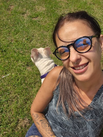 A woman is taking a selfie outdoors on a grassy area. She is wearing black-rimmed glasses and a black and white patterned tank top. She has tattoos on her arm. Next to her is a white dog wearing a purple harness. The scene appears to be a park or garden. The location is not identifiable as Bridlington, United Kingdom.