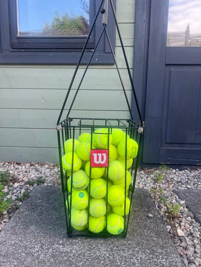 A black metal basket filled with numerous bright yellow tennis balls sits on a concrete surface next to a gravel path. The basket is full, with balls piled high and spilling over the sides slightly. One of the tennis balls has a green circular mark on it. A red rectangular tag with a white "W" logo is visible on the front of the basket. Behind the basket, a light green wooden-paneled wall is visible, and above that, a dark blue wooden section of a building, possibly a door or window frame. A reflection of trees and a sky can be seen in a dark-framed window to the left. The scene appears to be outdoors, in daylight, with bright sunshine illuminating the tennis balls. The setting suggests a tennis court or a practice area, likely in a residential or recreational space. There is no activity taking place, the basket of balls is stationary. The overall impression is one of readiness for a game or practice.