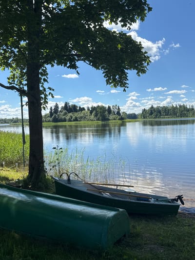 This is a photograph of two green boats on the shore of a lake in Lendži, Latvia. The boat in the foreground is partially visible, showing its green hull. The second boat, in the middle ground, is a smaller green rowboat with oars and an outboard motor. It is positioned near the water's edge, adjacent to tall reeds. A large tree with green leaves shades a portion of the scene on the left. The lake extends to the horizon, where a dense line of trees forms the background. The sky is blue with scattered white clouds, reflecting on the calm surface of the water. The overall environment suggests a tranquil, natural setting.