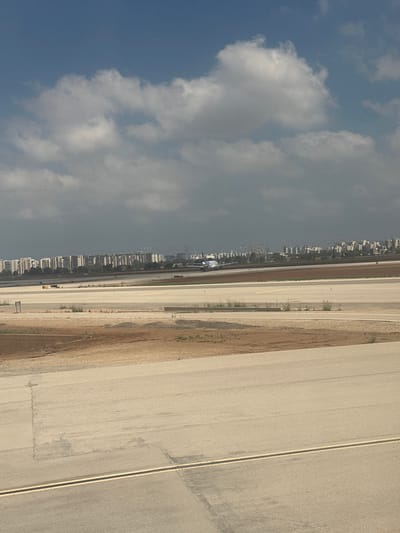 An airplane is on a runway at Ben Gurion International Airport in Yehud-Monosson, Israel. The sky is blue with scattered white clouds. In the background, a densely populated urban area with numerous buildings stretches across the horizon. The runway is made of asphalt and has yellow and white markings. The ground surrounding the runway is a mix of dry grass and dirt. The airplane is white with blue accents and is facing away from the viewer. It appears to be either taxiing or preparing for takeoff.
