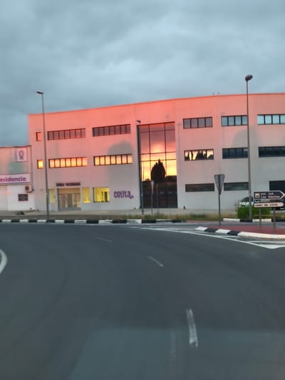The media file presents an outdoor scene featuring a modern commercial building, an asphalt road, and street furniture under an overcast sky. The time of day appears to be either sunrise or sunset, as indicated by the intense orange-pink light reflected prominently on the building's white facade and within many of its windows.
The main subject is a two-story, white building with numerous rectangular windows. A large section on the right side of the building features dark, highly reflective glass, mirroring the vivid orange sky and silhouettes of trees. The road in the foreground is paved with asphalt, showing white lane markings and a prominent curb with alternating black and white stripes, suggesting a roundabout or intersection. Two tall streetlights stand on either side of the road.
No people are visible, and the scene is static. The overall mood is quiet and atmospheric due to the dramatic natural light contrasting with the grey sky.
Visible text provides location and business cues:
* On the left side of the building, partially visible text reads "residencia" with a logo above it, possibly indicating a care home.
* Below this, a sign identifies a "clínica veterinaria" (veterinary clinic).
* Further right on the building, purple graffiti spells "ceuta".
* A brown road signpost on the right directs traffic towards "pol. ind. Los Vientos" (Industrial Park Los Vientos), "camí de Llíria" (Road to Llíria), and points to a "hotel." These place names are consistent with the provided context of Nàquera / Náquera, Spain.