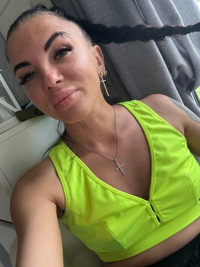 This media file features a close-up, portrait-style shot of a woman lying down, with the camera positioned to capture her face and upper body. She has dark hair in a braid, extensive eyelash extensions, and visible piercings on her ear and nose. She is wearing a bright neon yellow top, possibly a sports bra or bikini top, with a delicate silver necklace featuring a cross pendant. The background includes a textured grey fabric, likely upholstery from a couch or chair, and hints of a window with natural light suggesting daytime. The overall mood appears casual and relaxed. The specific location is identified as Южное Снт, Russia. There is no visible text in the image.