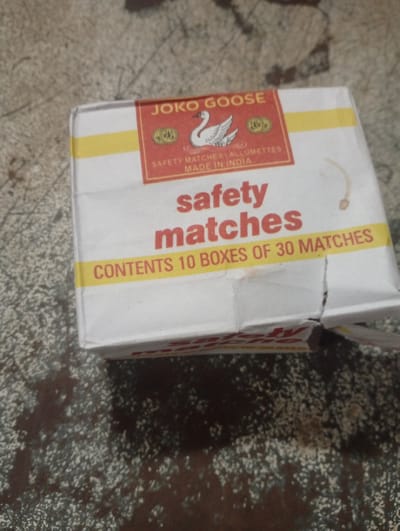 A box of safety matches is on a textured, speckled surface. The box is white with a red and yellow label. The label has the text "JOKO GOOSE" at the top, a picture of a swan, and smaller text that reads "SAFETY MATCHES ALLUMETTES MADE IN INDIA". Below this, in large red letters, are the words "safety matches". Further down, in yellow text, it says "CONTENTS 10 BOXES OF 30 MATCHES". The box appears to be slightly damaged on the right side. The textured surface is a mottled brown and grey. The image is taken from a slightly elevated angle, looking down. The location in Jalingo, Nigeria, is not discernible from the image.