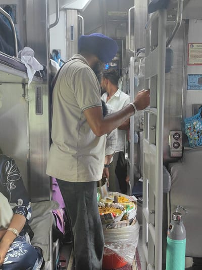 A man wearing a blue turban and a beige shirt is standing in a train compartment. He is holding a tray of food, which includes what appears to be fried snacks and some hard-boiled eggs. To his right, another man in a white shirt is visible. The train compartment is narrow, with bunk beds on either side. There is a blue mesh bag hanging on the wall, filled with various items. A turquoise and gray water bottle is also visible on the right side of the image. The environment appears to be a public transportation setting, possibly a train in India. The image does not provide any specific details about the city of Mandwa, India, or any events taking place there.