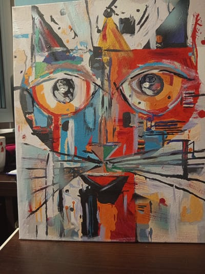 The image is a close-up, head-on view of an abstract painting of a cat's face on a canvas. The painting is characterized by bold, angular shapes and a vibrant, clashing color palette, featuring reds, oranges, blues, yellows, and blacks. The cat's eyes are large and prominent, rendered with concentric circles and detailed pupils. The whiskers are depicted as sharp, black lines. The background is a textured white canvas with splotches and drips of various colors, suggesting a dynamic and energetic artistic process. The painting is displayed on a wooden surface, possibly a table, and is slightly tilted. The overall impression is one of a stylized, perhaps even fragmented, representation of a feline subject. There is no indication of any specific location within the city of Чэрвеньскі сельскі Савет, Belarus.