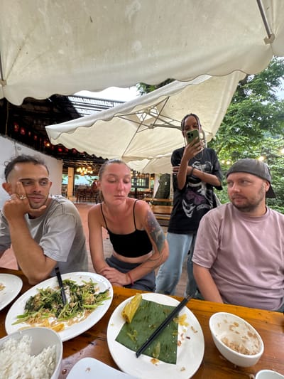 Four people are seated at a wooden table under a large white umbrella. The background appears to be an outdoor restaurant in Cổ Lũng Commune, Vietnam, with trees and a building visible. In the foreground, there are plates of food, including rice and stir-fried vegetables. One person, a woman with dark hair in braids, stands and takes a selfie with her phone. The other three individuals are seated, appearing to be engaged in conversation or observing their surroundings. The lighting suggests it is daytime, possibly overcast or shaded.