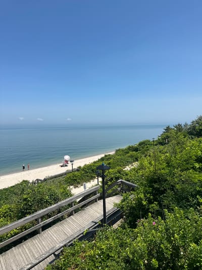 The image shows a view of a beach and the ocean from an elevated wooden boardwalk. The boardwalk curves down towards the sandy beach, which is lined with lush green bushes and trees. On the beach, a lifeguard chair with an umbrella is visible, and two people are standing near the water. The ocean is calm and extends to the horizon under a clear blue sky with a few wispy clouds. The scene appears to be located in the Town of Riverhead, United States of America, likely on the north shore of Long Island, which is known for its beaches and waterfront access. The time of day is daytime, with bright sunlight casting shadows.