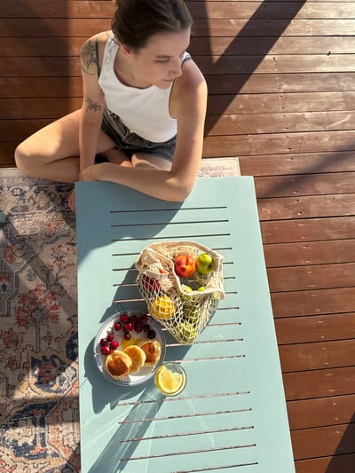 A medium shot captures a young woman in a white tank top and denim shorts sitting on a wooden deck, her legs crossed and arms resting on a light blue slatted table. She is looking down and to her right, away from the camera. The deck is bathed in dappled sunlight, creating strong shadows. On the table, to the woman's right, is a white mesh bag filled with green apples and a few other pieces of fruit, including what appear to be nectarines. To the left of the mesh bag, on a white plate with a blue rim, are three small pancakes, a cluster of cherries, and a slice of lemon in a clear glass. A patterned rug is visible on the deck to the left of the table. The environment suggests an outdoor setting, likely a patio or balcony. There is no indication of the city of Starokorsunskaya, Russia within the image itself.