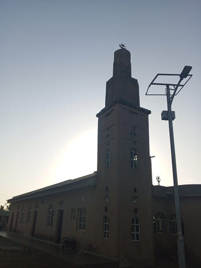 The image shows a mosque in Tawpo, Nigeria. The mosque is a single-story structure with a tall minaret. The minaret has a crescent moon and star on top. A street light is visible next to the mosque. The sky is light blue, and the sun is behind the building, creating a silhouette. The scene is quiet, with no visible people or activity.