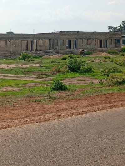 The image shows an exterior view of a long, rectangular building under construction in Shuwa, Nigeria. The building appears to be a single-story structure with an open roof and partially completed walls made of concrete blocks. Numerous doorways and window openings are visible along the facade, some with exposed wooden framing. The surrounding environment consists of a grassy, unkempt field with patches of dirt and scattered vegetation. In the foreground, a dirt road or path runs diagonally across the lower portion of the image, leading to a paved road. The sky is overcast with a few visible clouds. There are no people or vehicles visible in the scene.