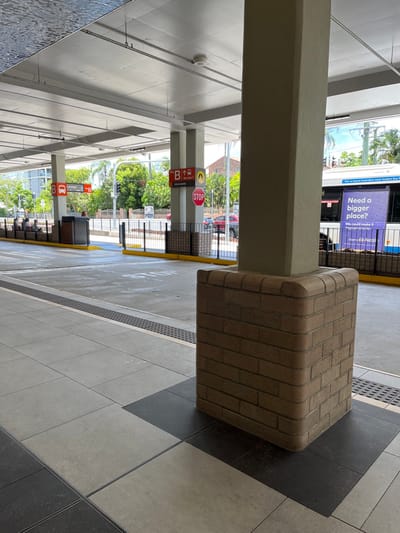 The image depicts an outdoor public transport platform, likely a bus stop or interchange in Indooroopilly, Australia, under bright daylight conditions. In the foreground on the right, a large, square pillar stands, featuring a base covered in light brown brickwork, while its upper section is plain beige. To the left and slightly further back, a narrower beige pillar holds multiple signs. These include a prominent red circular "STOP" sign and a red square sign marked with a white "B," a bus icon, and an upward arrow, indicating a specific bus bay or route. Another smaller sign on this pillar features a yellow circle with a black 'V' symbol.

In the background, a blue and white bus is partially visible, displaying an advertisement with the text "Need a bigger place?" and "Get on board Australia's most modern NSW bus." Beyond the platform, a street, metal fencing, and green foliage from trees are seen. The platform itself is paved with light grey and darker grey square tiles, arranged in a pattern, and features yellow tactile paving strips along its edge. The scene appears to be well-lit by the sun, casting shadows and indicating clear weather. An overhead canopy structure is visible, supported by the pillars. No discernible human activity or specific individuals are clearly visible in the frame.
