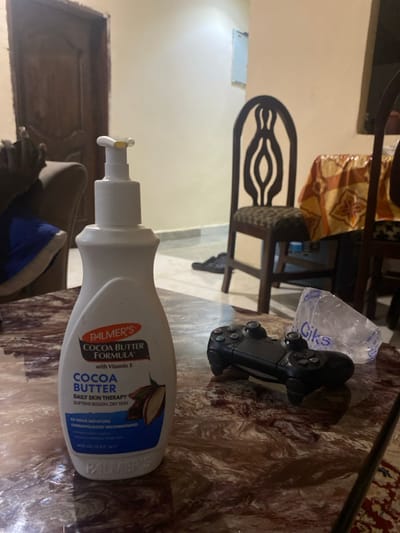 The image displays an indoor scene, likely a living or dining area, in Lakka, Sierra Leone. The primary focus is a white pump bottle of Palmer's Cocoa Butter Formula lotion, standing upright on a table with a marbled surface in the foreground. To the right of the lotion bottle, a black PlayStation controller is visible, alongside a clear, crumpled plastic bag with the word "Giks" printed on it.

In the background, various pieces of dark wooden furniture can be seen, including chairs. One chair's backrest features a distinctive, ornate design with elongated, eye-shaped cutouts. The walls are plain and light-colored, suggesting a simple interior decor. The floor also appears to be a light shade. A piece of upholstered furniture, possibly a sofa or armchair, is partially visible in the lower-left corner. No specific activity or event is taking place; the scene captures a static arrangement of everyday objects.

Notable text on the Palmer's lotion bottle includes "COCOA BUTTER FORMULA with Vitamin E", "COCOA BUTTER DAILY SKIN THERAPY SOFTENS ROUGH, DRY SKIN", "48 HOUR MOISTURE", "DERMATOLOGIST RECOMMENDED", and "400 ml / 13.5 fl. oz.". The word "Giks" is also clearly legible on the plastic bag. The lighting is consistent with an indoor setting, but does not indicate a specific time of day.