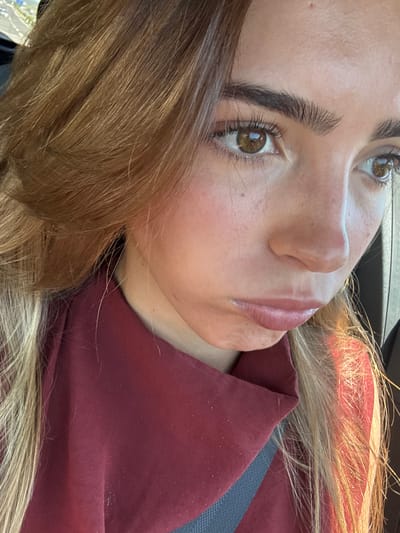 A close-up image depicts a young female individual seated within a vehicle. The individual's location is reported as Valado dos Frades, Portugal. The subject has long, light brown hair with blonde highlights, framing her face. Her large, brown eyes are directed to the viewer's right, showing visible long eyelashes and dark, thick eyebrows. Her nose is small, and her lips are closed in a slight pout. Skin texture with minor blemishes is discernible. She wears a dark red, collared garment. A dark gray seatbelt traverses her left shoulder. Natural light, likely sunlight, illuminates the right side of her face and hair. The background is blurred, consistent with a vehicle interior.
