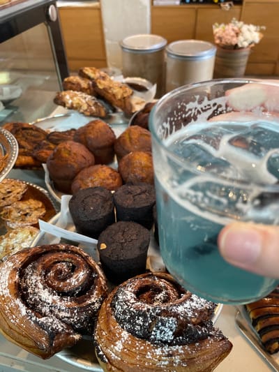 A variety of baked goods fill the frame, including cinnamon rolls dusted with powdered sugar, cylindrical dark chocolate cookies, and golden brown muffins. Glassware is also visible, creating a blurred, abstract effect over the pastries. Metal canisters stand in the background above a display case containing more almond pastries. The image is shot from a low angle, emphasizing the abundance and variety of the treats.