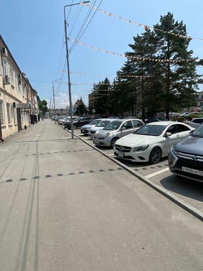 The image depicts a street scene in Spassk-Dal'nij, Russia. The perspective is from a low angle, looking up towards a clear blue sky and across a row of parked cars. The cars are lined up on a paved street, with white parking lines visible. In the foreground, the side of a building with multiple windows and air conditioning units can be seen. Overhead, wires are strung across the scene, and a line of small, colorful triangular flags is suspended between poles, suggesting a festive occasion or decoration.
The subjects in the image are primarily cars, with the suggestion of people inside some of them. The setting is an urban environment, likely a street or parking area in a town. The activity taking place is simply the presence of parked vehicles on a street on a sunny day.
Notable details include the bright sunlight, indicating it is daytime, likely mid-morning or afternoon. The clear sky suggests fair weather. The building in the foreground provides some architectural context, while the flags add a touch of color and occasion. Some text is visible on the license plates of the cars: one plate reads "P 105CK 125 RUS" and another appears to be "B 090 CB 125". The overall impression is a calm, ordinary street scene with hints of a celebration.