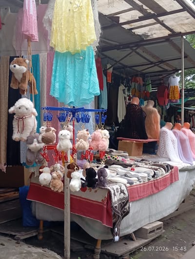 A stall displays handmade woolen items, including sweaters, socks, and plush toys, in Verkhnyaya Balkariya, Russia. The stall is covered by a corrugated metal roof supported by rusty metal beams. In the foreground, a white table is draped with a red and white patterned cloth, upon which various woolen socks and other knitted items are neatly arranged. Numerous small, plush toys resembling sheep and other animals hang from clips on a blue metal rack. Behind the table, several mannequins are dressed in crocheted sweaters in various colors, including black, peach, white, lavender, and pink. The background reveals more hanging garments and what appears to be the natural environment. The timestamp "26.07.2026 15:43" is visible in the lower right corner.