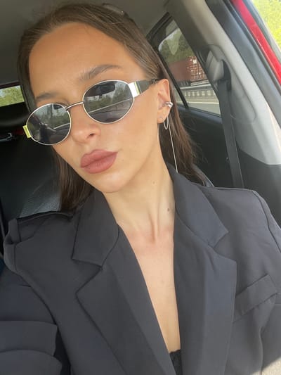 The image is a close-up, portrait-style photograph, rotated 90 degrees counter-clockwise, showing a person from the chest up inside a car. The individual, who appears to be female, is the primary subject, seen wearing large, round sunglasses with reflective lenses and a black blazer or jacket. She has dark hair, styled down, and her lips are painted with a muted, neutral-toned lipstick. Her visible right ear is adorned with multiple small ear cuffs and a long, delicate chain earring that dangles down.
The setting is the interior of a car, with parts of the headliner, a seat, and a red door frame visible in the background. Bright daylight streams in, creating highlights on the person's skin and strong reflections in the sunglasses. These reflections offer a glimpse of the outside environment, showing trees or foliage and what appears to be structures or buildings, suggesting a sunny outdoor day. The person is looking directly towards the camera, seemingly posing for a photograph. The overall impression is casual and stylish. There is no visible text in the image. This scene is set in De-Friz, Russia.