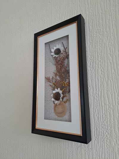 A framed shadow box arrangement of dried flowers, featuring two white daisy-like flowers, brown twigs, and small yellow flowers, is mounted on a textured, off-white wall. A miniature straw hat rests at the bottom of the arrangement. The shadow box has a black outer frame and a gold inner mat. The image is taken from a slightly upward angle, showing the side of the frame and the textured wall. No people or animals are visible. The scene is indoors, likely a home. The location is not specified in the image.