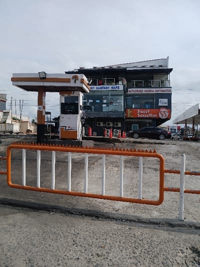 The image shows a gas station in Way, Nigeria, located at latitude: 6.429, longitude: 3.468. The scene is a gas station with a closed gate. The gas station is a concrete structure with a canopy over the pumps. There are three pumps, two of which are visible. The pumps are marked with the text "RIT SANITARY WAPE AUTHORISED NIGERIA DISTRIBUTOR" and "Sweet Sensation". There are no people in the scene, but there is a parked black car in front of the gas station. There are orange and white metal barricades extending from the pumps to the opposite side of the driveway. The ground is gray and paved. The sky is a bright overcast grey. The image was taken during the day. The image was taken in a horizontal orientation. The image is likely taken from a slightly elevated angle.